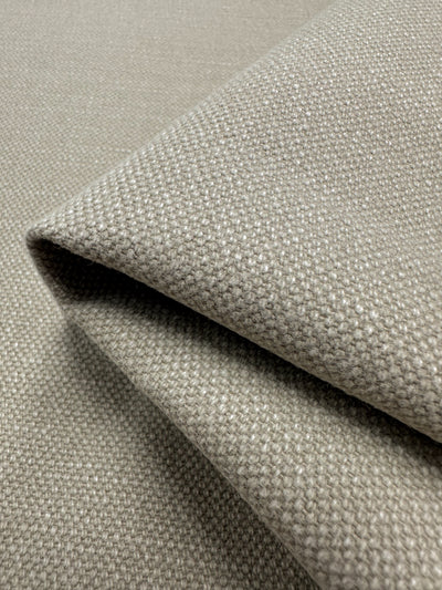 A close-up of a folded piece of the Canvas Linen Denim - Peyote from Super Cheap Fabrics, measuring 140cm. The fabric's heavyweight and textured surface in a neutral beige color resembles upholstery material, with visible fibers forming a subtle grid-like design. Gentle shadows are cast by the edges of the folds, accentuating its woven charm.