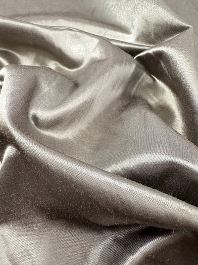 Close-up of Heavy Weight Satin from Super Cheap Fabrics, in a smooth, shiny Dune Metallic silver-gray with soft folds and a reflective surface. This 145cm upholstery material showcases a silky texture, offering a luxurious and elegant appearance.