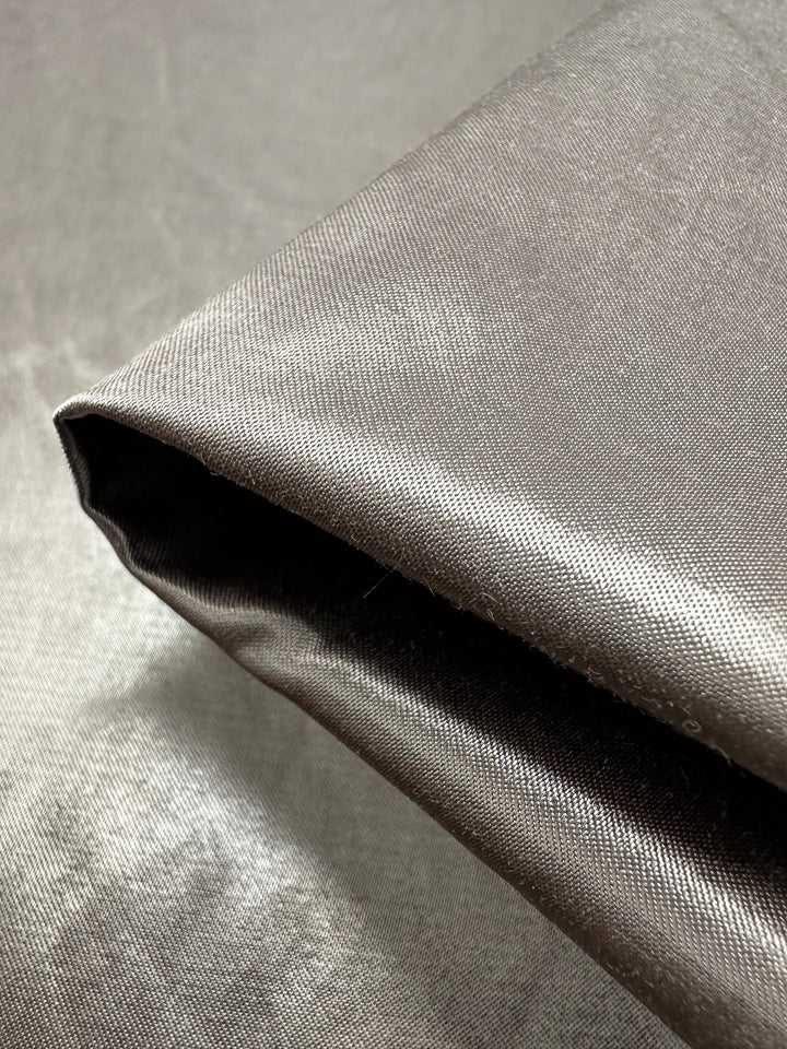 Close-up of a folded piece of Super Cheap Fabrics' Heavy Weight Satin - Dune Metallic - 145cm, with its dark gray hue and smooth texture that captures light, revealing subtle reflections on its lustrous surface—perfect for luxurious upholstery projects.