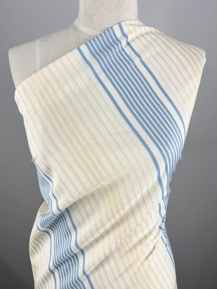 A mannequin draped in light-colored, *Super Cheap Fabrics' GOTS Cotton - Buttercream Stripe* fabric with vertical beige and blue stripes. The organic cotton fabric, measuring 160cm wide, wraps over one shoulder, highlighting the striped pattern. The background is a plain grey, emphasizing the eco-friendly fashion statement.