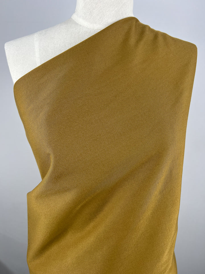 A mannequin is elegantly draped in a cumin-colored fabric named Gabardine Suiting - Cumin - 140cm from the brand Super Cheap Fabrics, covering the shoulder and chest area. This medium to heavy weight cotton fabric exhibits a smooth texture and appears soft, with delicate folds and drapes. The plain light gray background contrasts beautifully with the warm tone of the fabric.