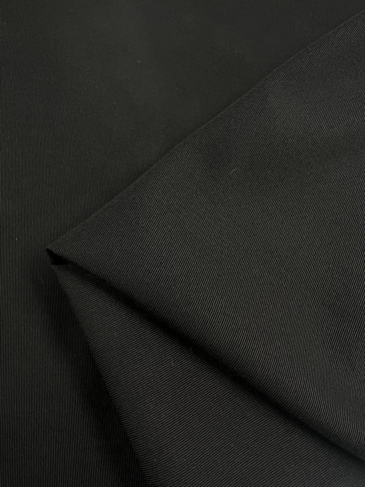 An image of Super Cheap Fabrics' Gabardine Suiting in black, a medium to heavy weight cotton fabric measuring 140cm wide, featuring a subtle ridge pattern. The fabric is folded over itself, creating layered and overlapping sections. The lighting highlights the texture and details of the material.