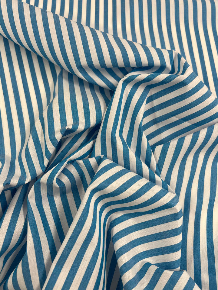 A close-up of the Super Cheap Fabrics' Cotton Lawn in Vivid Blue - 150cm showcases its lightweight cotton material, adorned with blue and white vertical stripes. The fabric is creased and folded, creating a textured appearance that enhances the visual complexity and depth of the textile—perfectly suited for warmer weather.