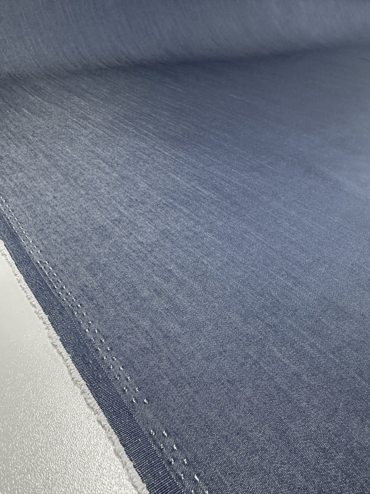 A close-up of a large piece of Super Cheap Fabrics' Chambray - Denim - 150cm highlights its smooth texture and light blue color. The edge of this fabric, which resembles denim, is visible with a slightly frayed seam running along the side.