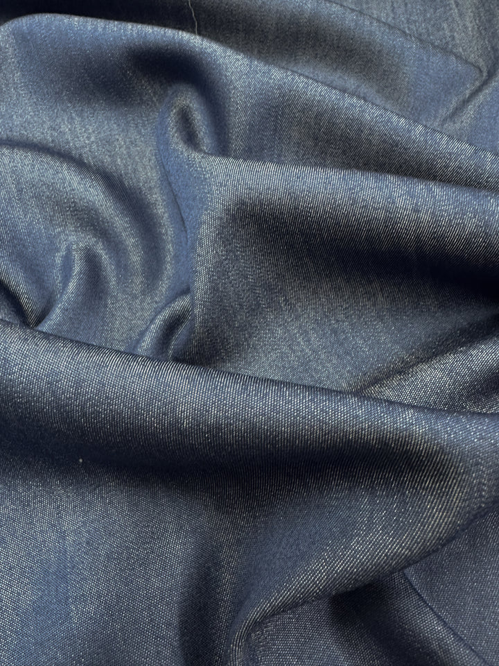 A close-up view reveals the Super Cheap Fabrics Chambray - Denim - 150cm, showcasing its smooth texture and visible weave patterns. This dark blue, lightweight fabric is gently folded, forming soft, flowing creases with subtle highlights and shadows.