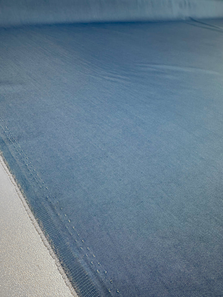 A close-up of the Chambray - Blissful Blue - 150cm fabric by Super Cheap Fabrics, neatly laid out on a flat surface. The smooth texture features visible stitching along the edges, with this serene blue fabric extending into the background and gradually fading out of focus.