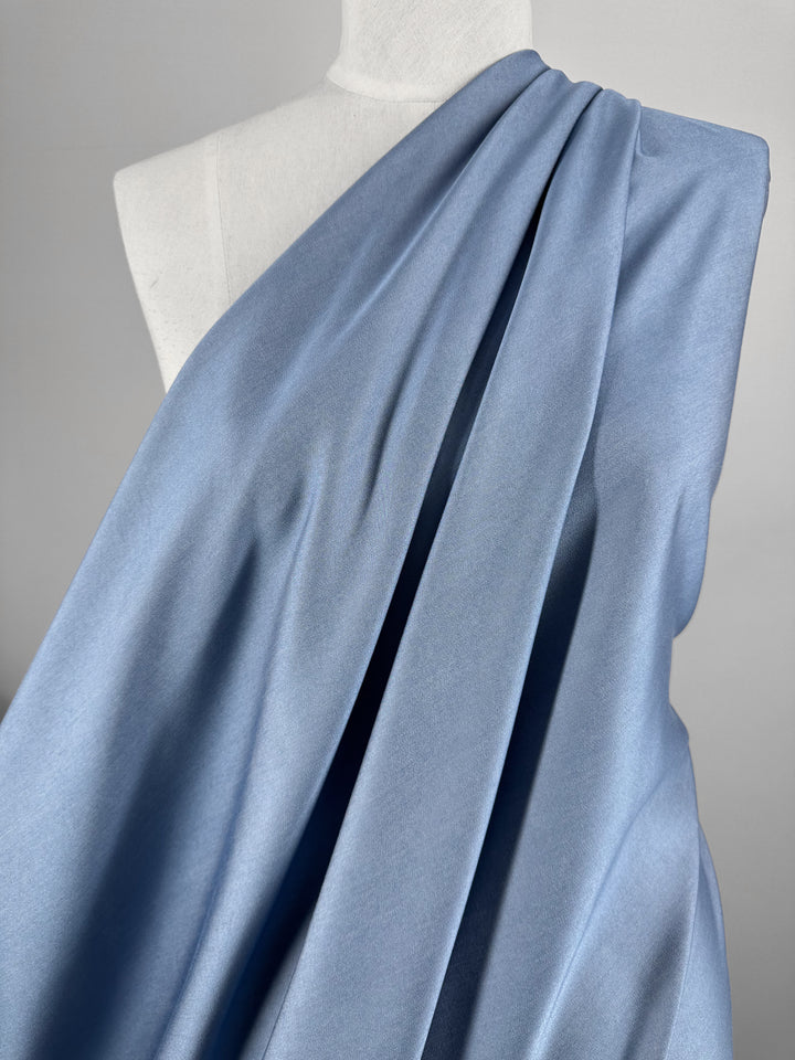 A mannequin draped in the Chambray - Blissful Blue fabric from Super Cheap Fabrics resembles a toga style. This lightweight material gently cascades over one shoulder, creating smooth folds and shadows.