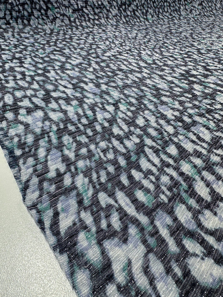 Close-up of the Metallix Pleat - Ocean Leopard fabric by Super Cheap Fabrics. This lightweight polyester features a textured pattern in navy, blue, green, and white tones. The design resembles abstract, scattered brush strokes against a darker background, creating a dynamic and flowing appearance. Width: 140cm.