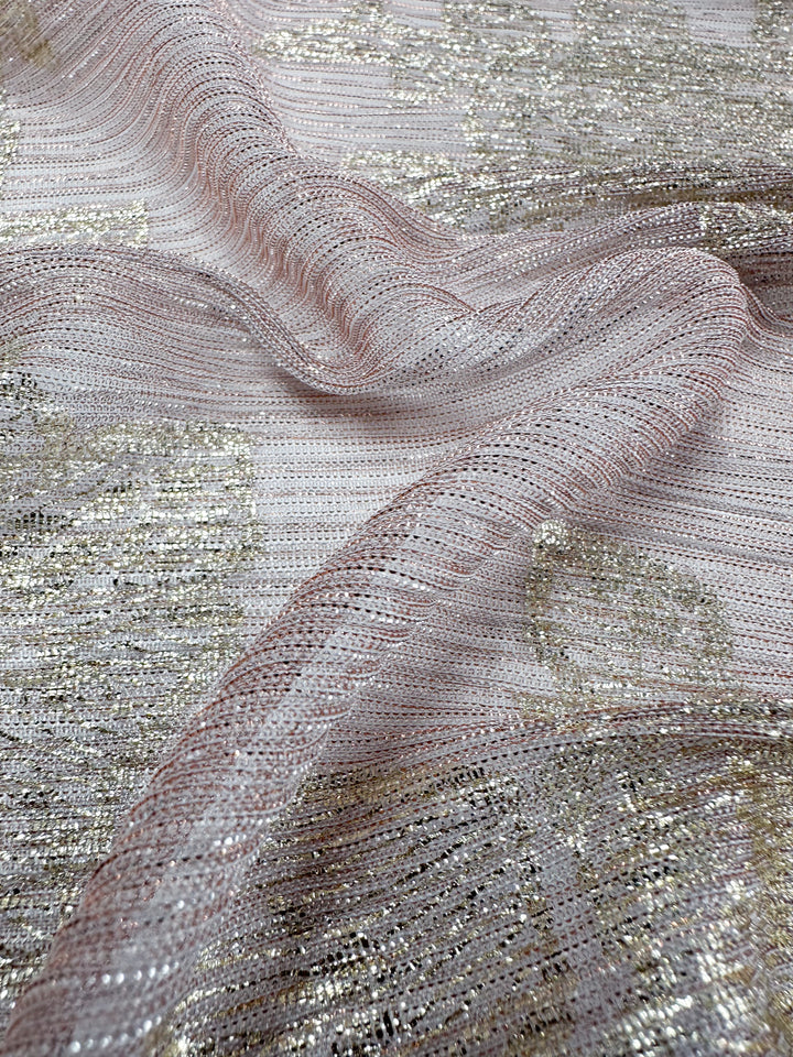 A close-up of the Metallix Pleat - Gold Leaf - 140cm polyester fabric from Super Cheap Fabrics reveals its light pink base, intricately interwoven with metallic silver and gold threads. The lightweight material has a soft, shimmery sheen with gentle folds that enhance its delicate appearance.