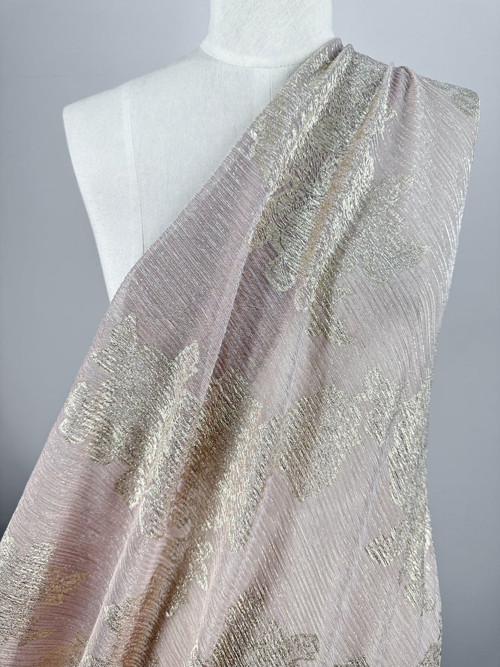 A draped mannequin is showcasing the Metallix Pleat - Gold Leaf fabric from Super Cheap Fabrics. This light-weight, luxurious material features a baby pink polyester backdrop adorned with an intricate, textured floral pattern in shimmering gold threads, highlighting its exquisite design details.