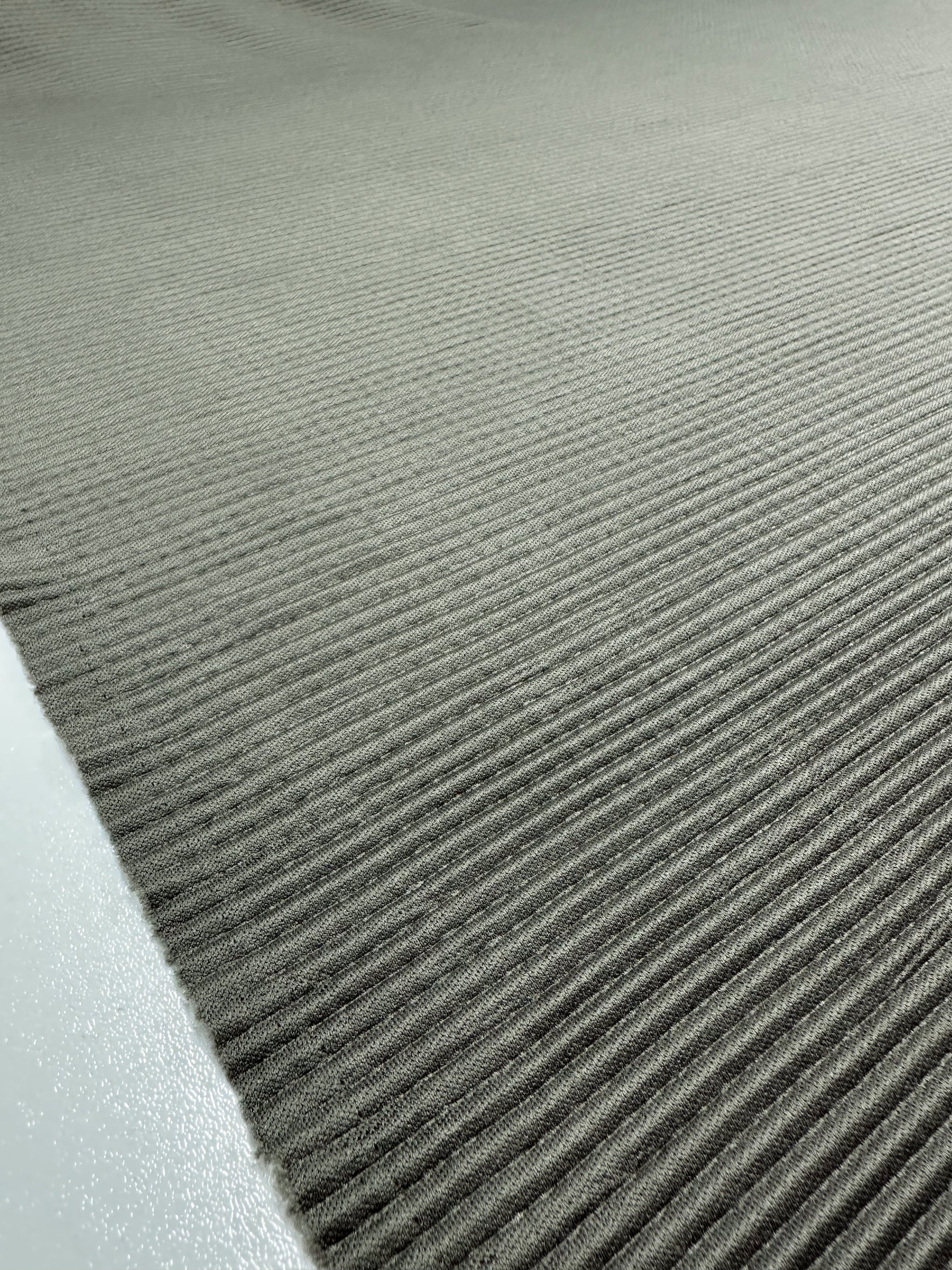 The close-up shows the ribbed, textured surface of Pleat - Pewter - 140cm by Super Cheap Fabrics. The gray polyester fabric fills the frame with evenly spaced parallel ridges and a contrasting matte finish on the smoother left edge.