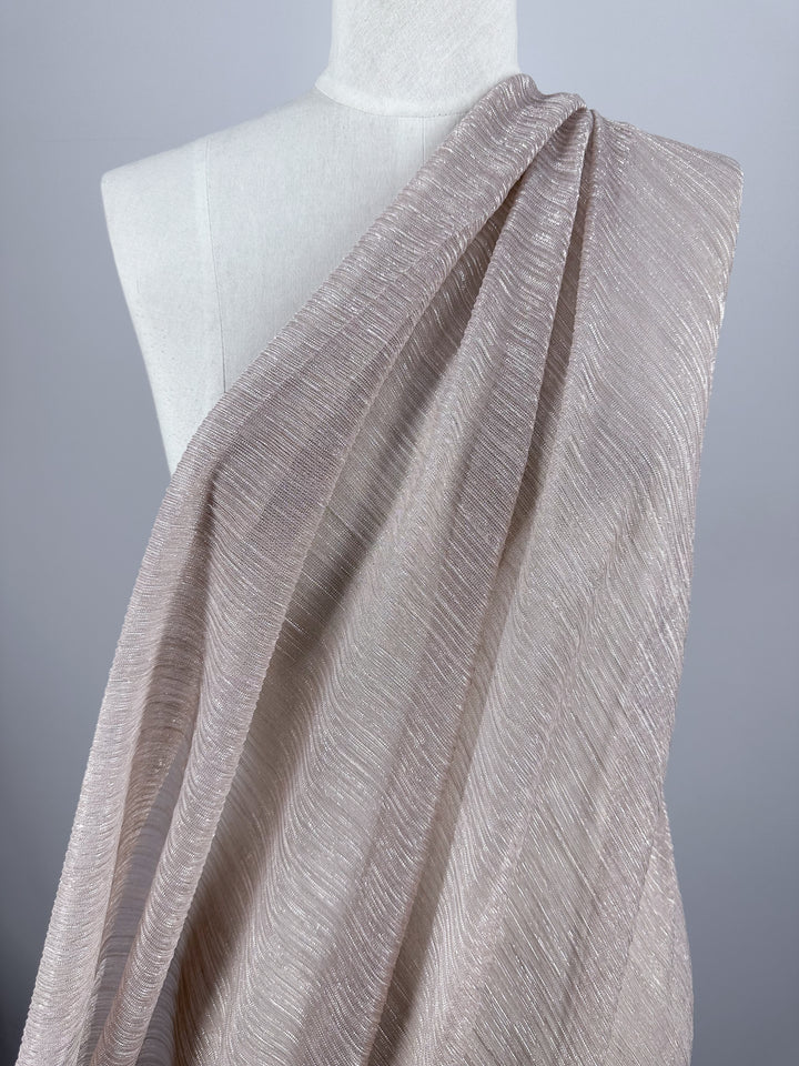 The Metallix Pleat - Blush - 140cm from Super Cheap Fabrics is elegantly draped over a mannequin, highlighting its subtle shimmer and soft pleats in light beige polyester. This lightweight fabric is beautifully presented against a simple, neutral gray backdrop.