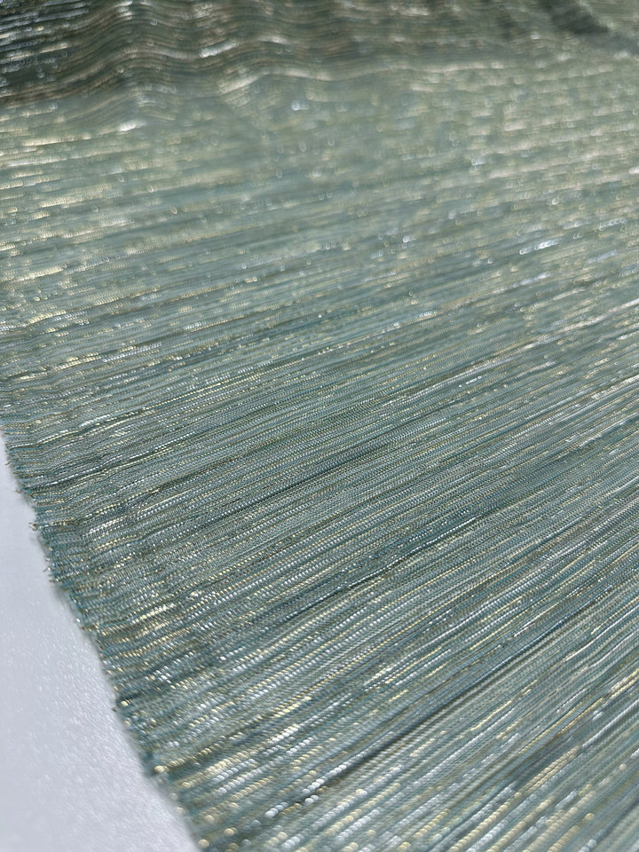 A detailed view of the Metallix Pleat - Mermaid Glitter - 140cm from Super Cheap Fabrics displays a textured polyester fabric interwoven with shimmering metallic gold threads in a gentle, light blue hue. The threads create an undulating pattern that reflects light for a glossy finish. The surface looks smooth and finely woven, providing a lightweight sensation.