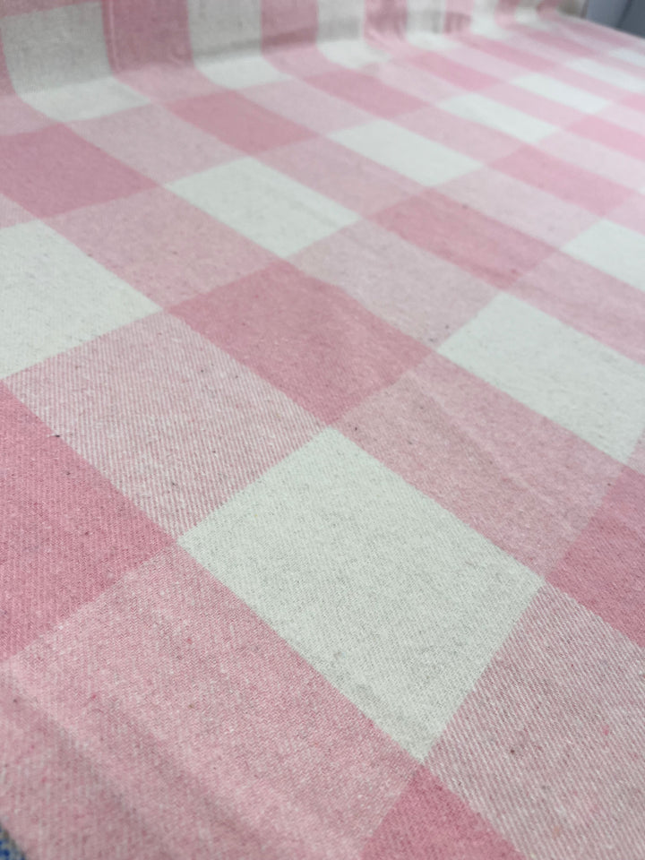 A "Tickled Pink" tablecloth from the brand Super Cheap Fabrics, featuring large pink and white checkered squares, is draped over a table. The flannel plaid fabric has a heavy weight and a woven texture, providing both durability and a delightful appearance. The 100% polyester composition ensures long-lasting quality for any occasion.