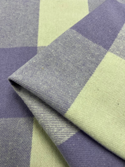 Close-up view of folded heavy-weight Flannel Plaid fabric by Super Cheap Fabrics, featuring a large checkered pattern in light green and viola hues. The texture appears soft and woven, and the edges are sharply creased. Width measures 148cm.