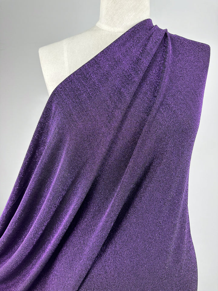 A mannequin elegantly displays the smooth texture of Super Cheap Fabrics' "Metallic Lycra - Disco Plum - 160cm." This shimmery purple fabric is artfully arranged in a flowing, designer dress style over one shoulder, set against a plain white background.