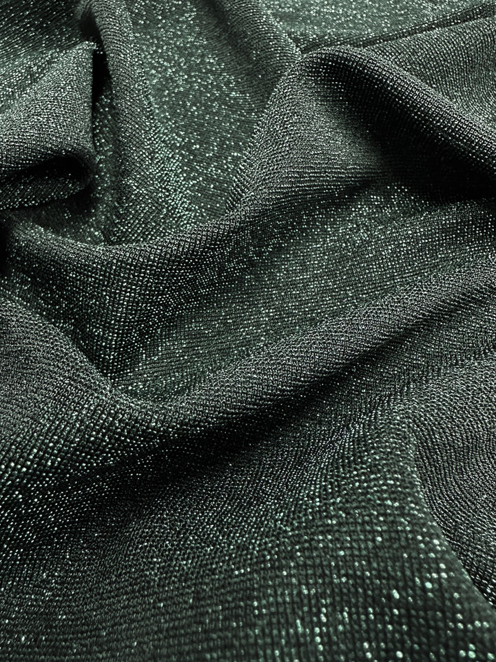 The close-up of the Metallic Lycra from Super Cheap Fabrics reveals a wavelike pattern created by reflective specks. The smooth and glossy texture, accentuated by light highlighting the sparkling details, is reminiscent of elegant dresses and is featured in their chic green jacket with a 170cm width.