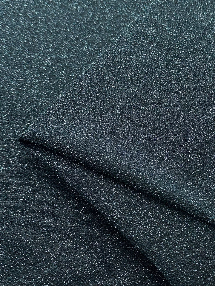 Close-up of the sparkling Aqua Fuzz Metallic Lycra fabric by Super Cheap Fabrics, showcasing its dark gray hue and shimmering texture. The folded section at the bottom right corner highlights its glittery surface, making it perfect for crafting stunning dresses.