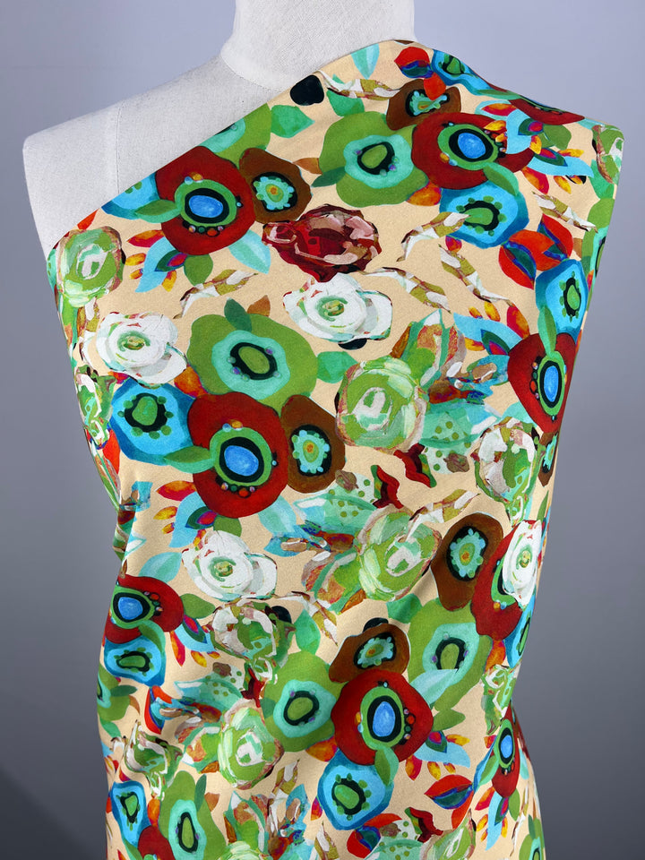 A mannequin is draped with the versatile Designer Rayon - Eloise fabric from Super Cheap Fabrics, featuring a colorful and vibrant floral pattern. The design includes large, abstract flowers in shades of green, red, blue, and white against a beige background.