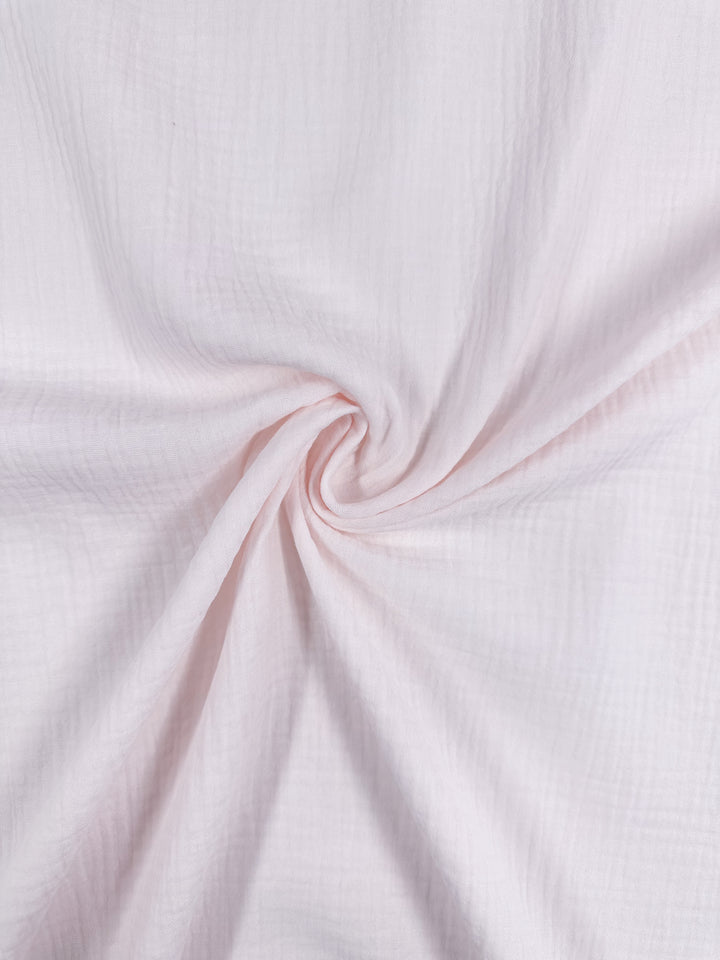 A close-up of the Muslin - Slightly Pink - 135cm fabric from Super Cheap Fabrics reveals its soft, light pink hue and textured surface. The twisted organic cotton forms a spiral pattern in the center, showcasing its delicate and airy quality.