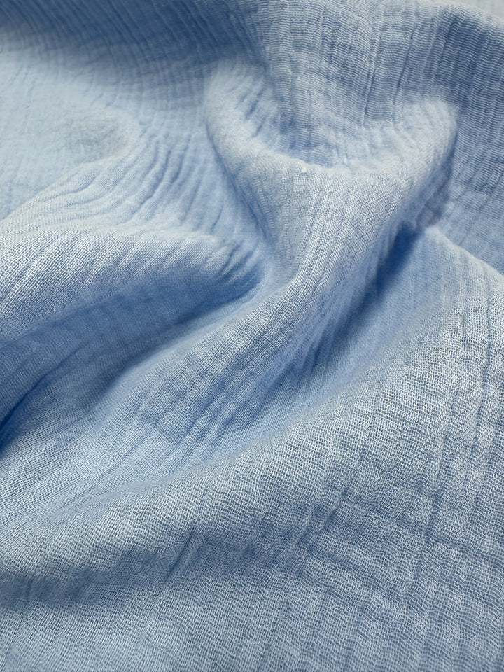 A close-up view of Super Cheap Fabrics' "Muslin - Serenity - 135cm" in light blue reveals a soft, crumpled texture. This organic fabric looks lightweight and gauzy, showcasing subtle grid-like patterns and gentle folds.