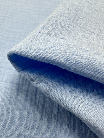 Close-up of folded Serenity muslin fabric by Super Cheap Fabrics, in a light blue hue that highlights its smooth yet gently crinkled texture. This organic Muslin - Serenity - 135cm material is arranged to showcase the intricate variations in its weave, emphasizing its durable and reusable nature.