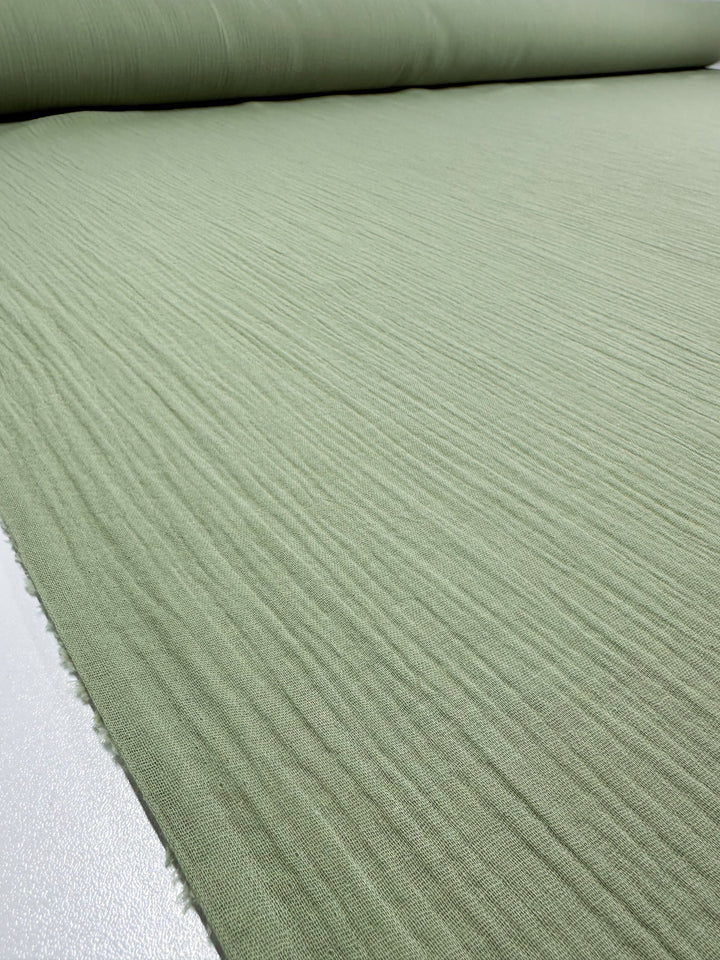 A close-up of a roll of Muslin - Laurel Green - 135cm fabric by Super Cheap Fabrics shows its textured surface. The organic fabric is partially unrolled on a flat surface, showcasing its linear pattern.