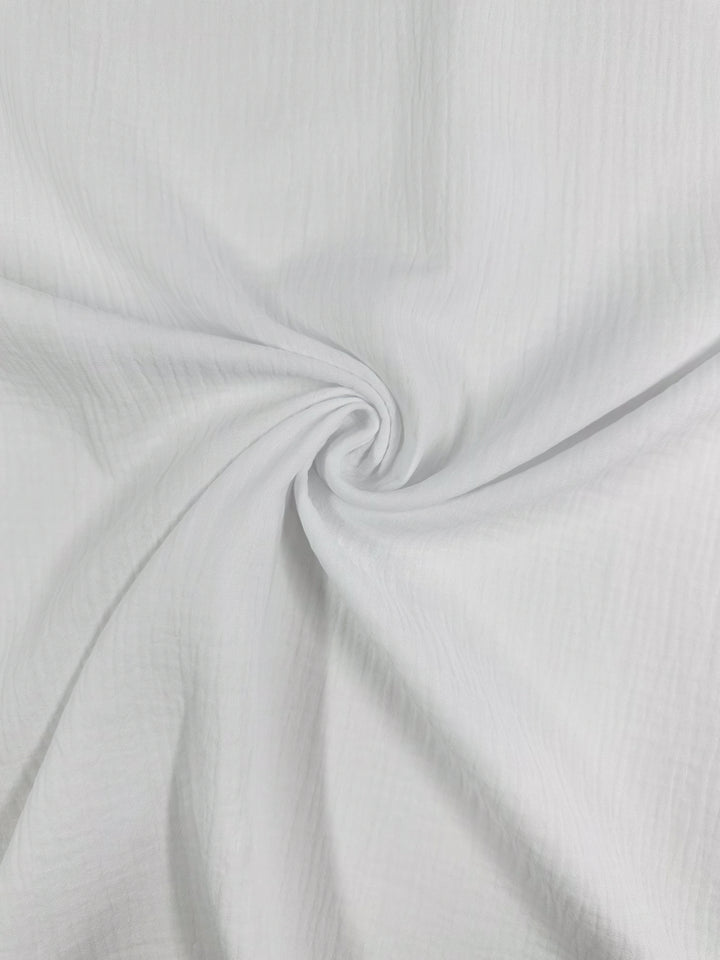 A close-up of the Snow White Muslin fabric from Super Cheap Fabrics, featuring a twisted swirl pattern in the center. The texture is soft and slightly crinkled, with gentle folds radiating outward, highlighting the natural elegance of this organic cloth, which measures 135cm.