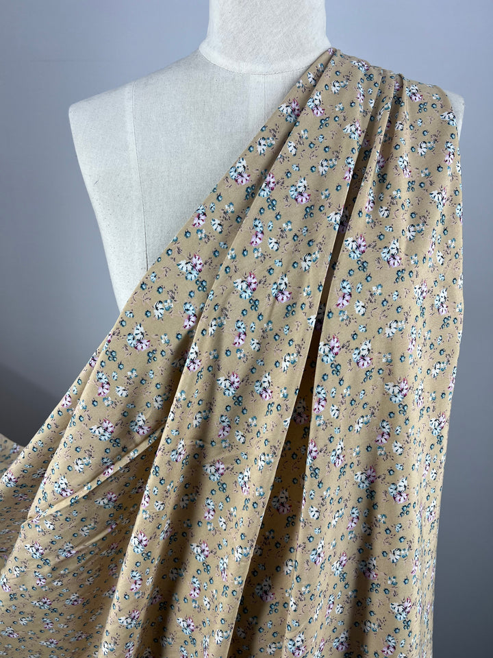 A mannequin draped with Super Cheap Fabrics' Printed Rayon - Pansy - 145cm, showcasing a delicate pattern of small pink, white, and green flowers on beige fabric. The versatile choice extends from one shoulder across the torso, highlighting its elegant drape and texture against a plain light-colored background.