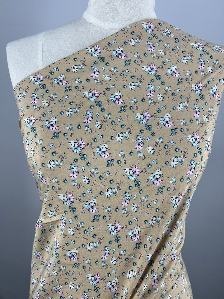 The mannequin is draped in "Printed Rayon - Pansy - 145cm" from Super Cheap Fabrics. This beige fabric features a small floral pattern with white, blue, and pink pansies accompanied by green leaves. Styled to cover one shoulder, it showcases a simple and elegant design.