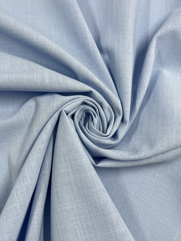 A soft, light blue Linen Blend - Dutch Canal fabric from Super Cheap Fabrics, featuring a subtle textured pattern, is gathered and twisted in the center to create a spiral effect. The material appears smooth and drapes gracefully. The image accentuates the fabric's color and texture, evoking the serene hues of a Dutch canal.