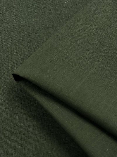 A close-up of the Linen Blend - Four Leaf Clover fabric by Super Cheap Fabrics, with a folded edge. The dark green fabric is textured with a fine weave and appears to be smooth and soft. The image focuses on the material, highlighting its color, texture, and lightweight drape.