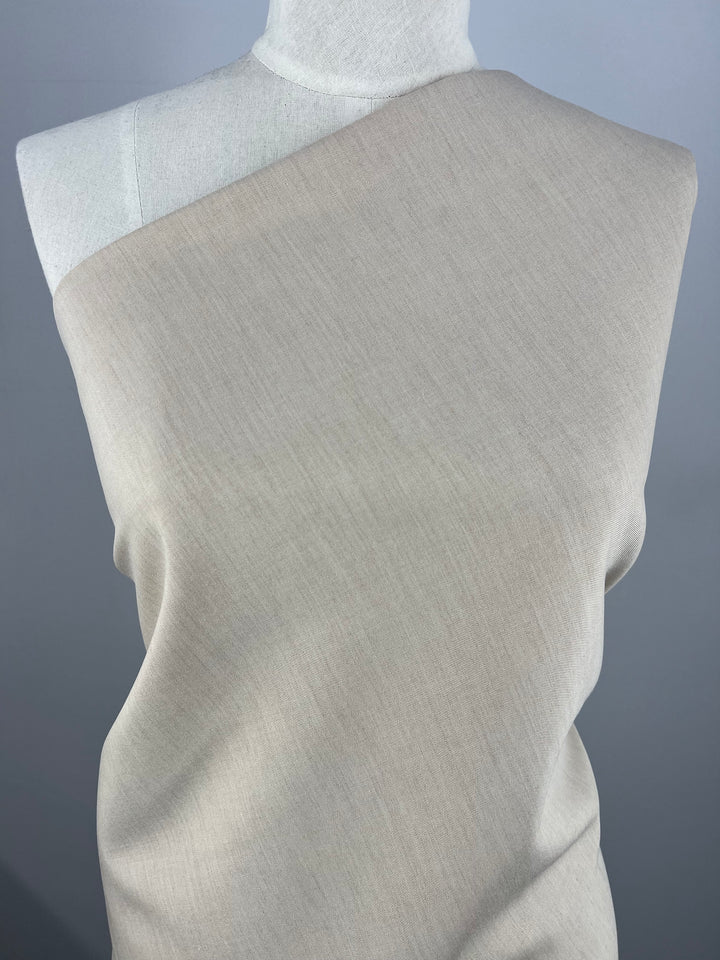 A mannequin draped with *Designer Suiting - Sandy - 150cm* from Super Cheap Fabrics, set against a plain gray background. This medium to heavy weight suiting polyester fabric possesses a subtle texture and falls smoothly over the mannequin's form.