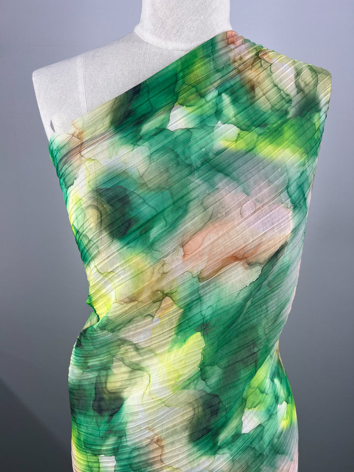 A mannequin is adorned with "Textured Tencel - Mossy - 140cm" from Super Cheap Fabrics, featuring a single-shoulder design in an abstract, watercolor-like pattern with shades of green, yellow, pink, and white. The pleated texture enhances its lush, vibrant appearance while offering unparalleled softness and sustainability.