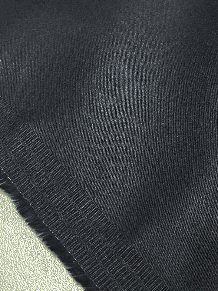 A close-up of Plain Sateen - Navy by Super Cheap Fabrics shows its textured surface with a frayed edge on a light green material. The cotton edge features horizontal patterns contrasting with the smoothness of the lighter sateen fabric beneath.