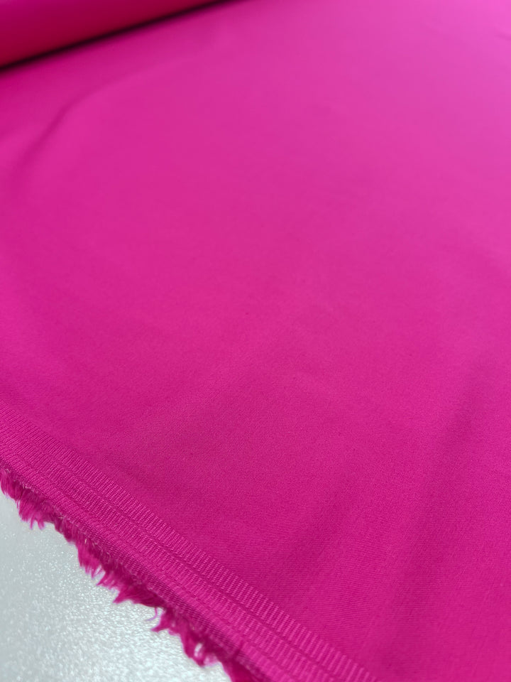 A close-up of Super Cheap Fabrics' Plain Sateen in hot pink, featuring a slight texture and frayed edge on the left, is draped over a white surface to highlight its vibrant hue.