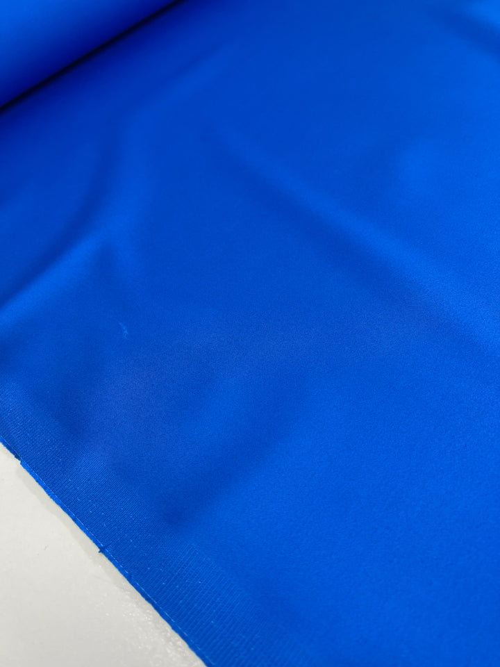 A close-up of Super Cheap Fabrics' Plain Sateen in royal blue, sized at 140 cm, reveals its smooth texture and vibrant folds. This bright fabric, ideal for clothing and décor, pops against a light background.