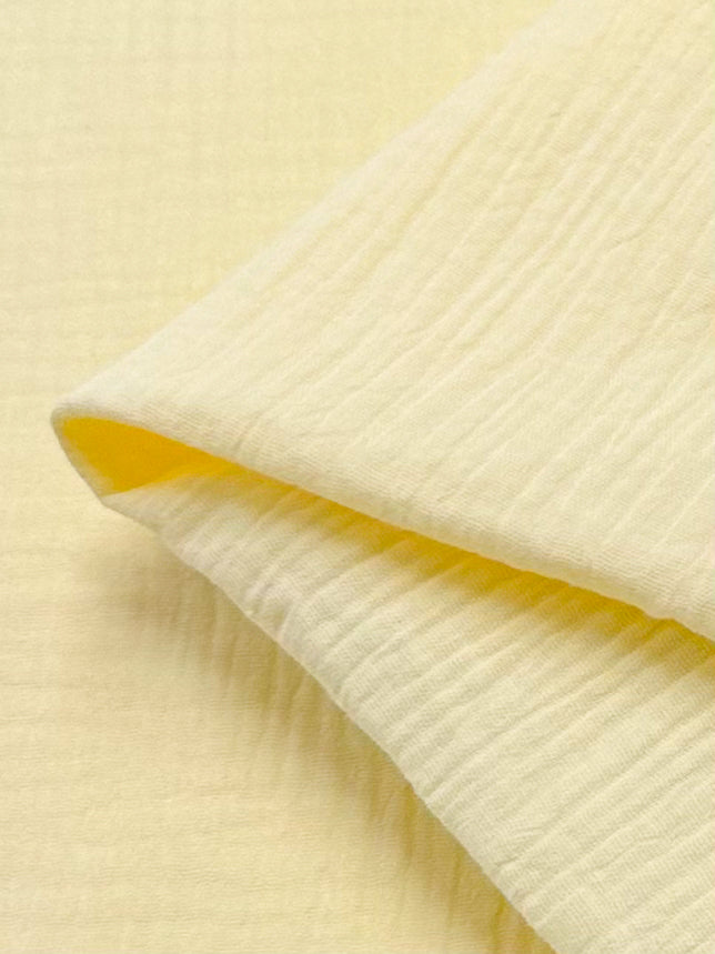 A close-up view showcases a folded piece of soft, textured muslin in pastel yellow from Super Cheap Fabrics, set against a matching background. The organic fabric is lightweight and gauzy, with dimensions of 135 cm.