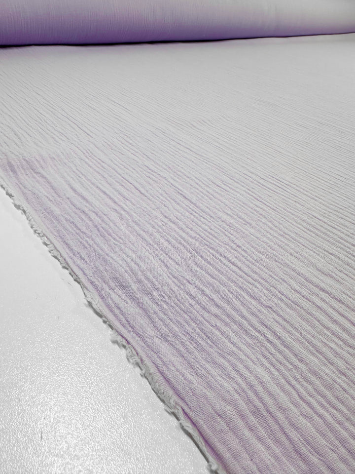 A close-up view of a large roll of Muslin - Lilac Breeze - 137cm from Super Cheap Fabrics, featuring a textured surface, laid out on a clean white table. The fabric's edges are slightly frayed, showcasing its ribbed texture.