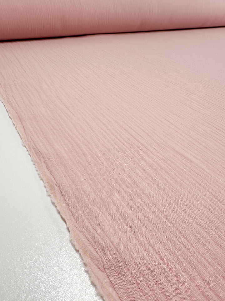 A close-up view of a roll of light pink Muslin - Lotus - 135cm fabric by Super Cheap Fabrics. The organic material features a ribbed texture and is spread out on a white surface.