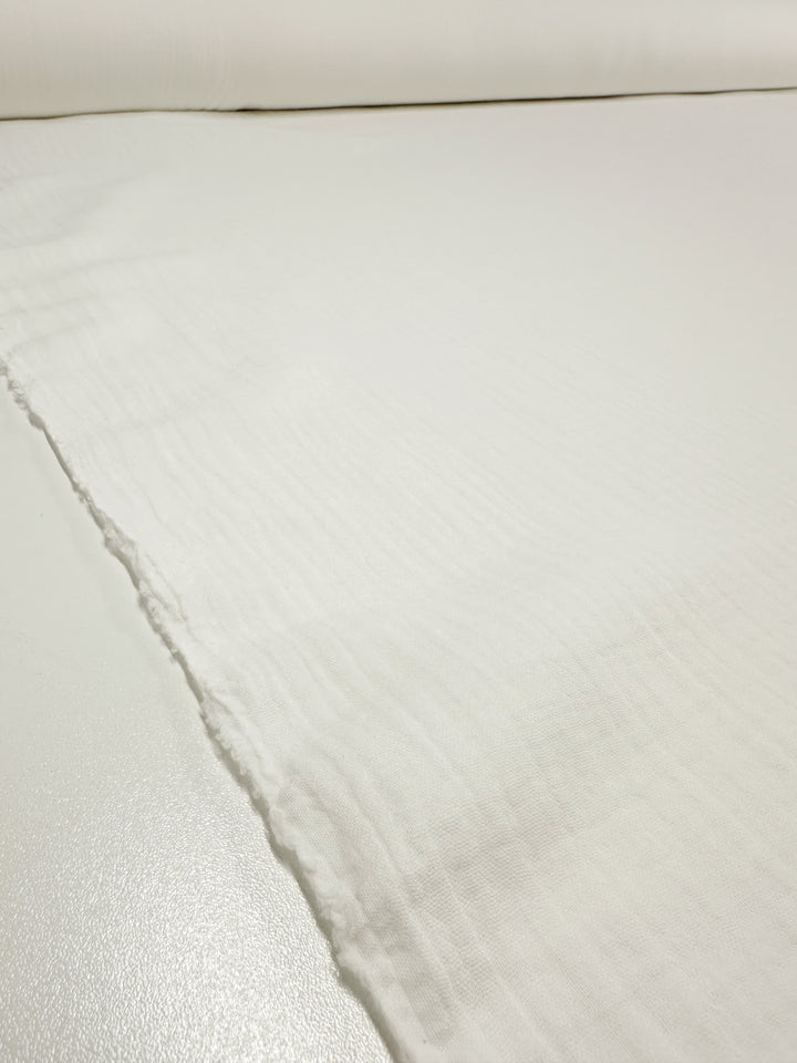 A detailed close-up of the Super Cheap Fabrics Muslin in Ivory, 135cm wide, showcases its lightweight and slightly textured surface. The fabric is spread out flat with a pristine, uncut edge visible, while the lighting accentuates its fine and smooth texture.