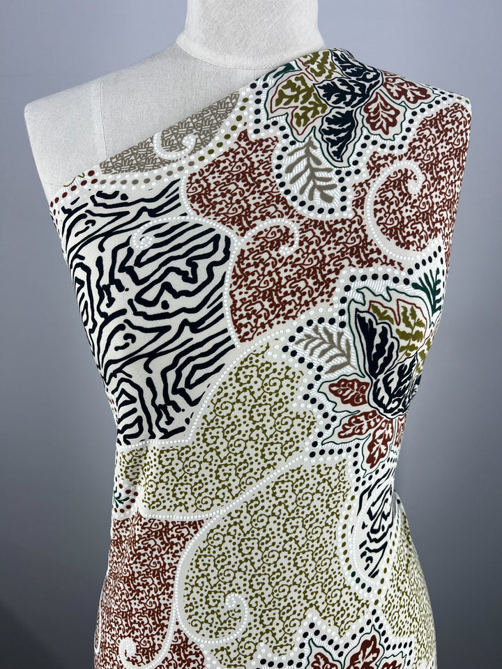 Displayed on the mannequin is an asymmetrical dress with a one-shoulder design, crafted from Super Cheap Fabrics' medium-weight Textured Lycra - Outline, featuring a vibrant pattern of leaves, swirls, and zebra-like stripes in black, red, green, and beige against a white background.