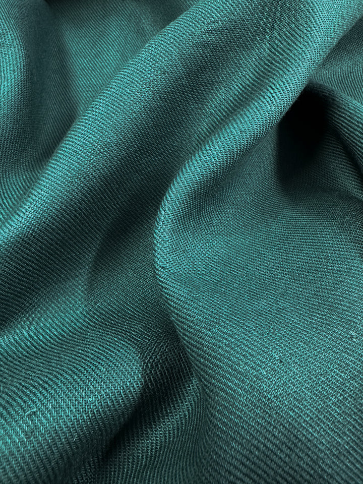 A close-up view reveals the texture and wavy folds of the "Pure Washed Linen - Jasper - 140cm" by Super Cheap Fabrics. This 100% linen material showcases a soft, smooth appearance with a subtly reflective surface that captures light in varying shades of teal and green.