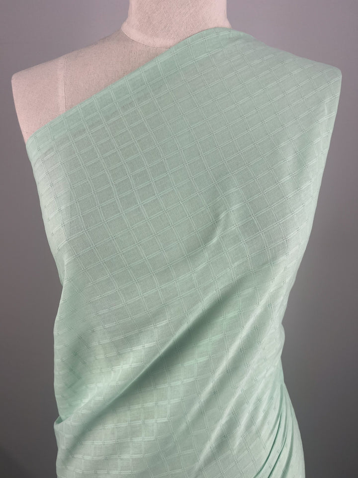 A mannequin draped in Super Cheap Fabrics' "Textured Cotton - Honeydew," a light mint green, extra lightweight cotton fabric, displays a subtle square grid pattern. The 100% cotton fibers are wrapped around the mannequin's torso, showcasing the fabric's texture and light sheen against a plain gray background.