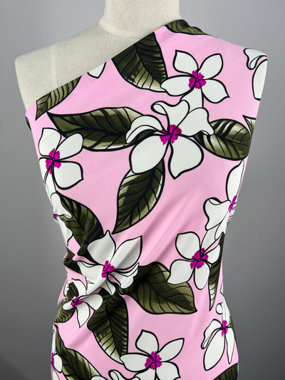 A close-up of a folded piece of Super Cheap Fabrics' Printed Nylon Lycra - Paraplu - 145cm draped over a mannequin. The high-quality print features large white flowers, green leaves, and pink accents on a light pink background. The mannequin is positioned against a plain gray backdrop.