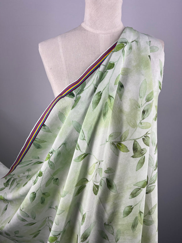 The Designer Cotton - EUCAS - 150cm from Super Cheap Fabrics is a lightweight cotton fabric in light green, adorned with a pattern of leaves. The fabric features a border with rainbow stripes, making it perfect for luxury sewing projects. The background is plain light gray in color.