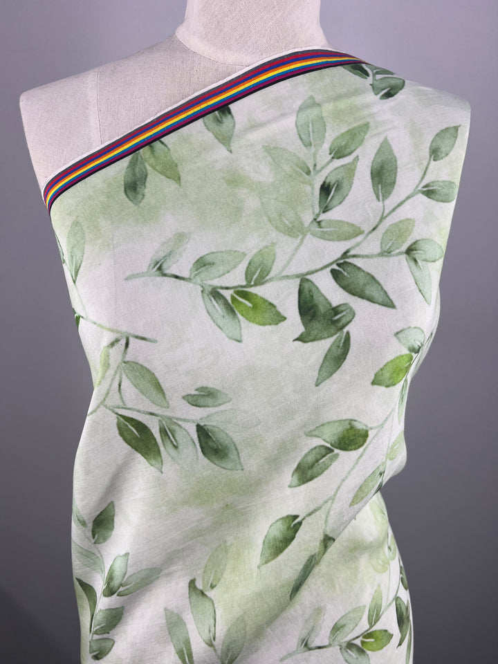 A mannequin is draped in the lightweight "Designer Cotton - EUCAS - 150cm" from Super Cheap Fabrics. This cotton fabric, decorated with a green leafy pattern, features a multicolored striped border near the top and has a neutral gray background that accentuates its vibrant design.