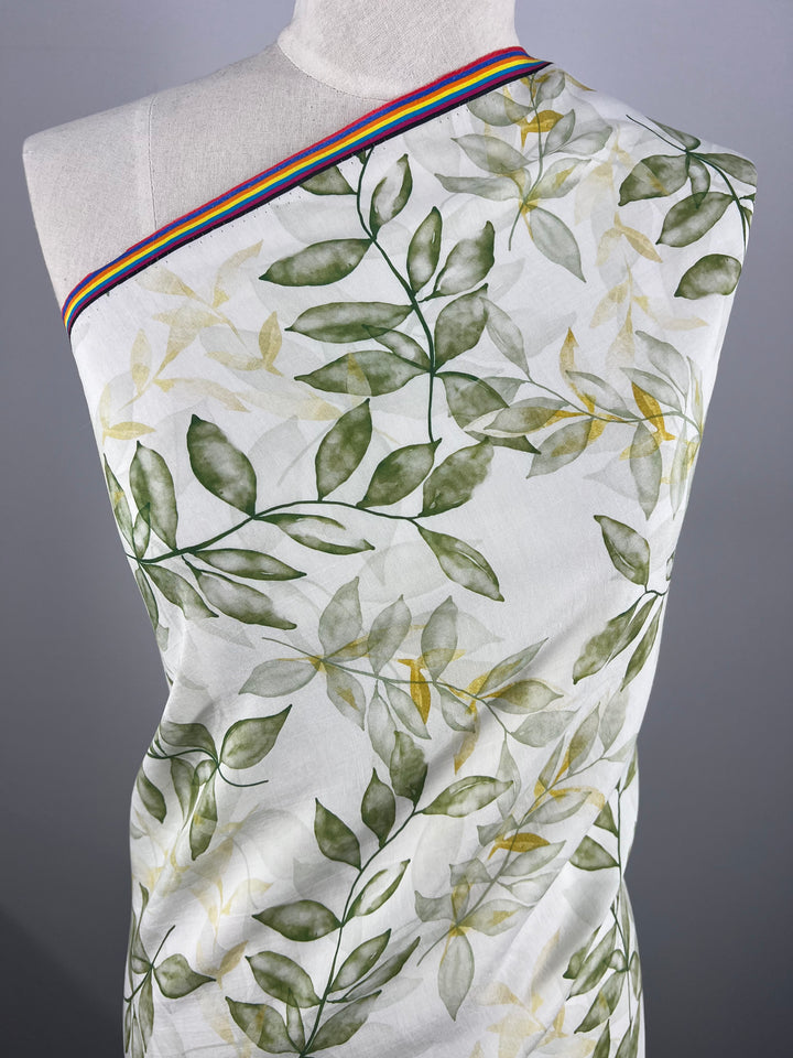 A mannequin draped in a luxury sewing project featuring Super Cheap Fabrics' Designer Cotton - Gully - 150cm, a lightweight cotton fabric with a green leaf pattern. The fabric is secured with a striped ribbon showcasing rainbow colors along the top edge. The background is plain and gray.