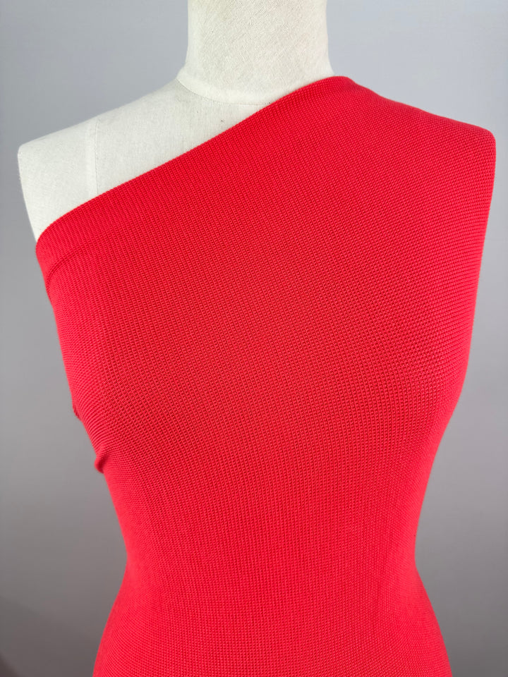 A white mannequin dressed in a vibrant red, one-shoulder, ribbed knit top against a plain gray background. The Honeycomb Knit - Watermelon - 158cm from Super Cheap Fabrics is form-fitting with a seamless design and subtle honeycomb fabric texture, enhancing the three-dimensional effect and highlighting the shoulder detail.