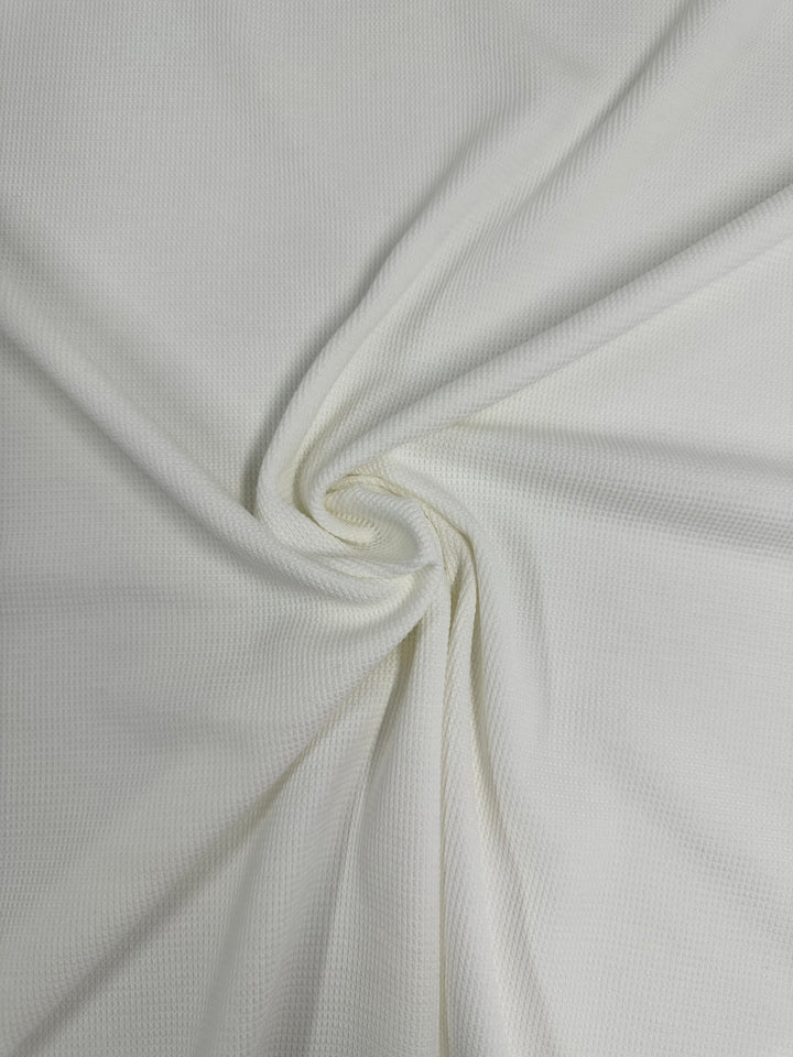 A textured white Honeycomb Knit - Vanilla - 158cm from Super Cheap Fabrics is depicted. The fabric is arranged in a swirl, creating a visually appealing spiral effect at the center. The material appears soft and pliable, with gentle folds radiating outward from the center of the spiral.